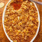 A casserole dish with a crispy golden-brown topping, possibly a gratin or baked dish, with a spoon inside and scattered potato chips around on a wooden surface.
