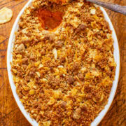 A casserole dish with a crispy golden-brown topping, possibly a gratin or baked dish, with a spoon inside and scattered potato chips around on a wooden surface.