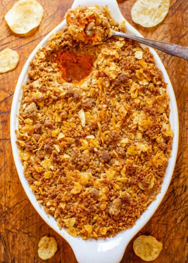 A casserole dish with a crispy golden-brown topping, possibly a gratin or baked dish, with a spoon inside and scattered potato chips around on a wooden surface.