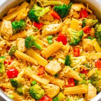 A pot of stir-fried noodles with chicken, broccoli, baby corn, and red bell peppers.