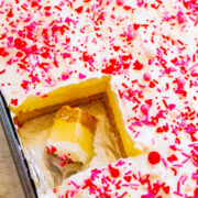 A tray of vanilla sheet cake with white frosting and red heart-shaped sprinkles, one piece partially removed.
