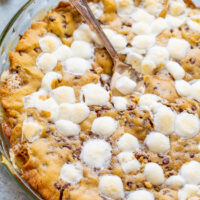 A freshly baked cookie pie with melted marshmallows on top, served in a glass dish with a spoon.