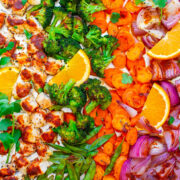 A colorful assortment of roasted vegetables and chicken on a baking sheet, including carrots, broccoli, red onions, orange slices, and herbs.