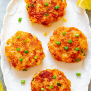 Four golden-brown seafood cakes garnished with green onions on a white plate, with lemon wedges and dipping sauce on the side.