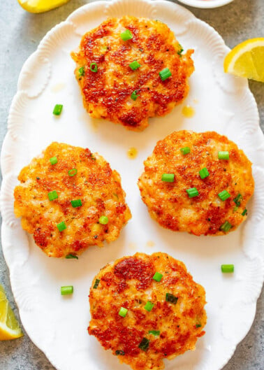 Four golden-brown seafood cakes garnished with green onions on a white plate, with lemon wedges and dipping sauce on the side.