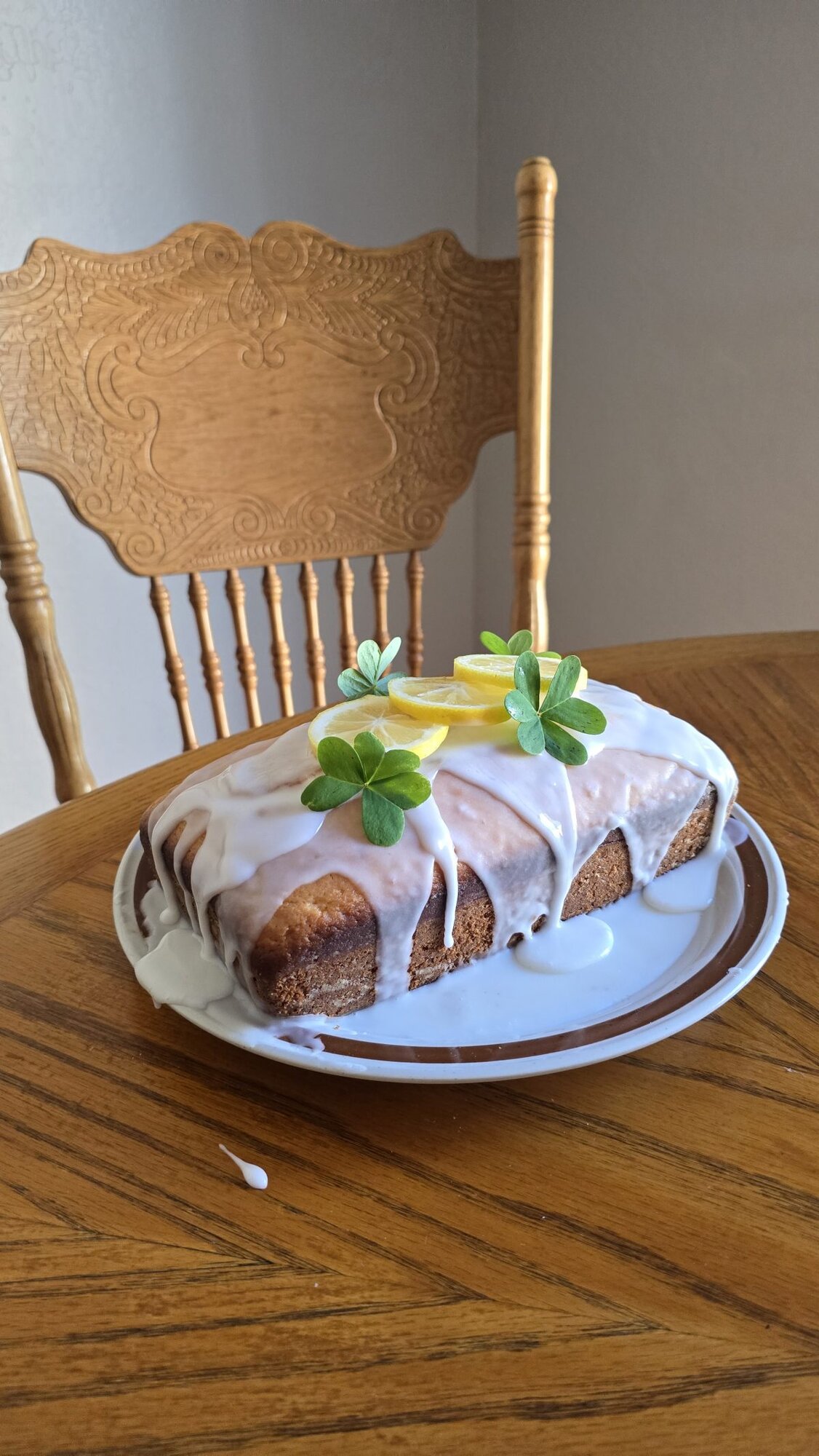 A loaf cake with white icing, topped with lemon slices and green leaves, sits on a white plate on a wooden table with a carved wooden chair in the background.
