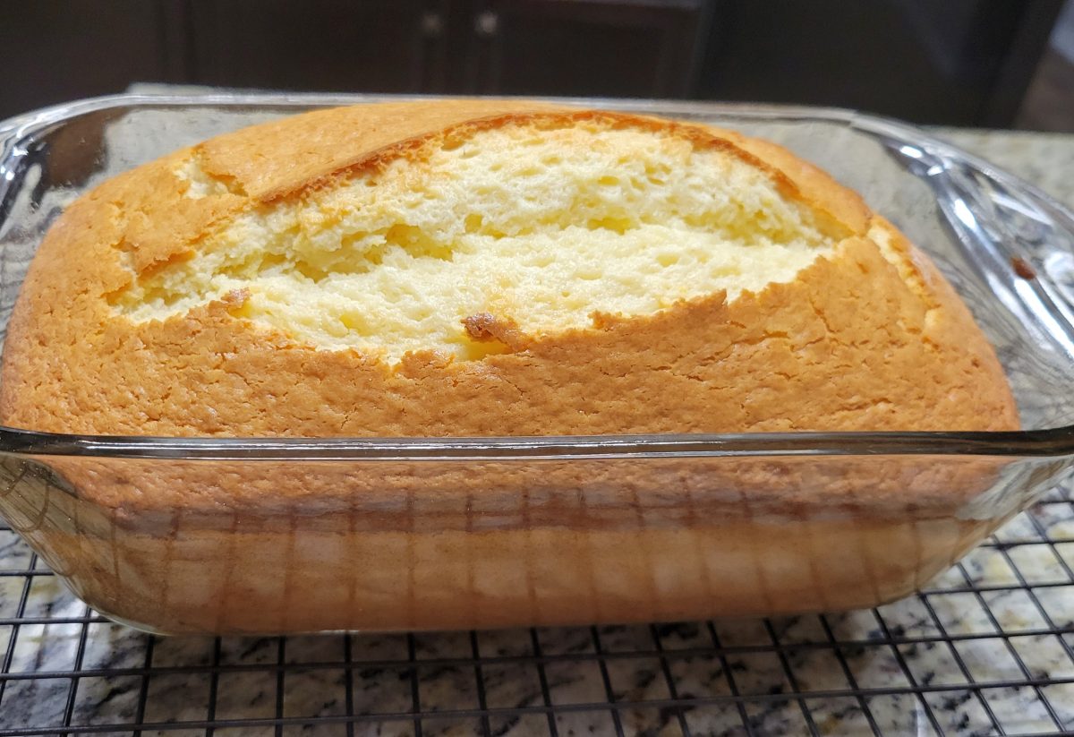 A loaf of golden-brown pound cake in a glass baking dish, resting on a wire cooling rack.