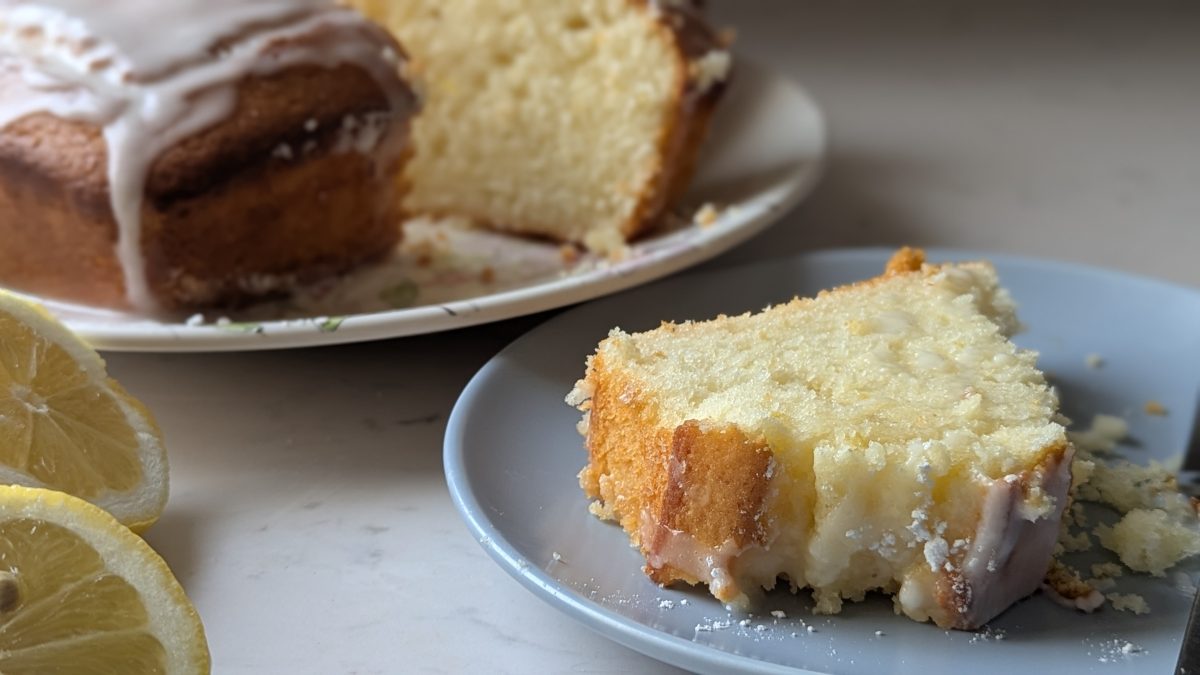 A slice of lemon pound cake with icing sits on a blue plate, with the rest of the cake and lemon halves in the background.