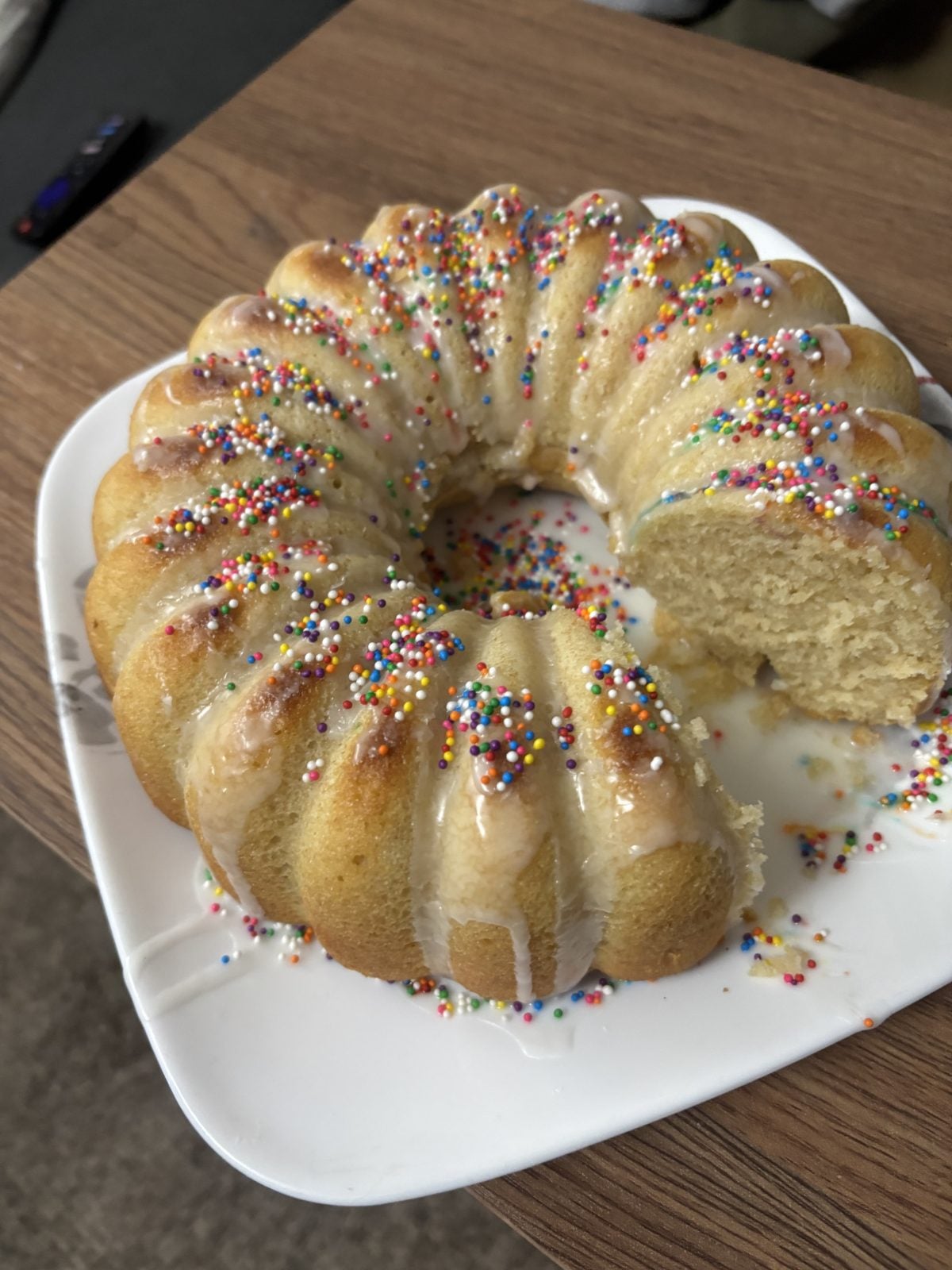 A round bundt cake with white icing and colorful sprinkles sits on a white plate, with one slice removed.