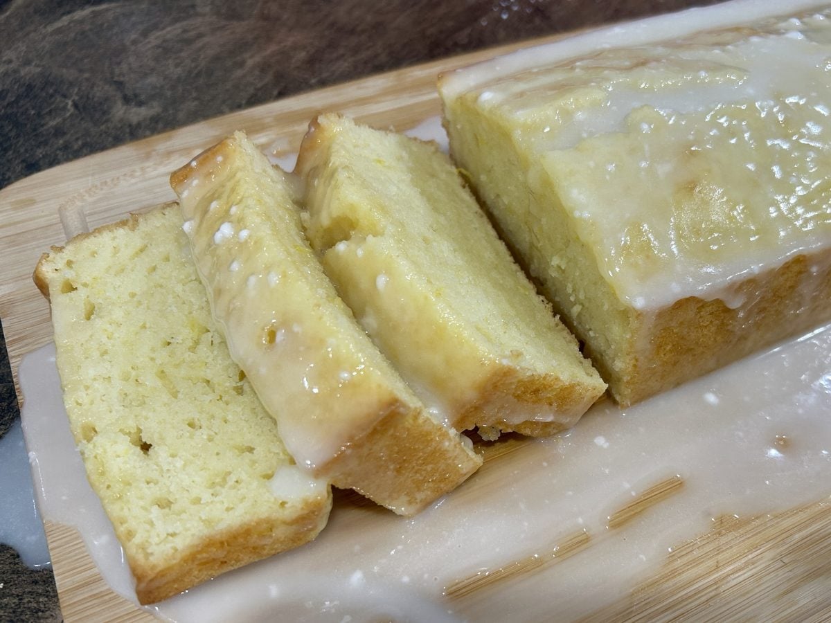 A loaf of glazed lemon bread on a wooden board, partially sliced, with icing dripped onto the surface.