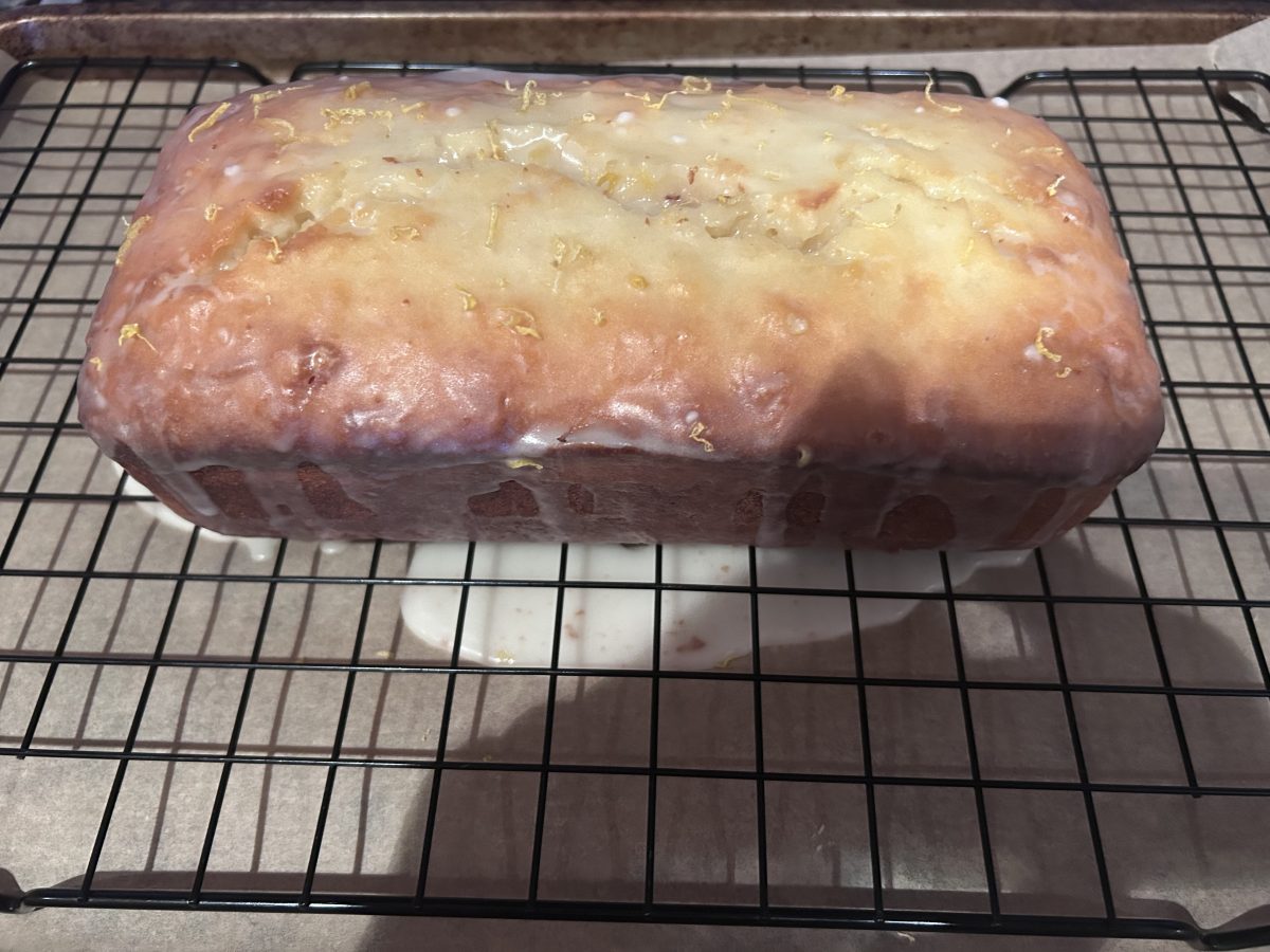 A glazed loaf cake with lemon zest sits on a cooling rack, with some glaze dripping onto the parchment-lined surface below.
