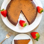 A chocolate cake dusted with cocoa powder, garnished with strawberries, with one slice served on a separate plate.