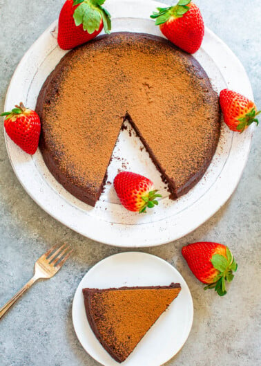 A chocolate cake dusted with cocoa powder, garnished with strawberries, with one slice served on a separate plate.
