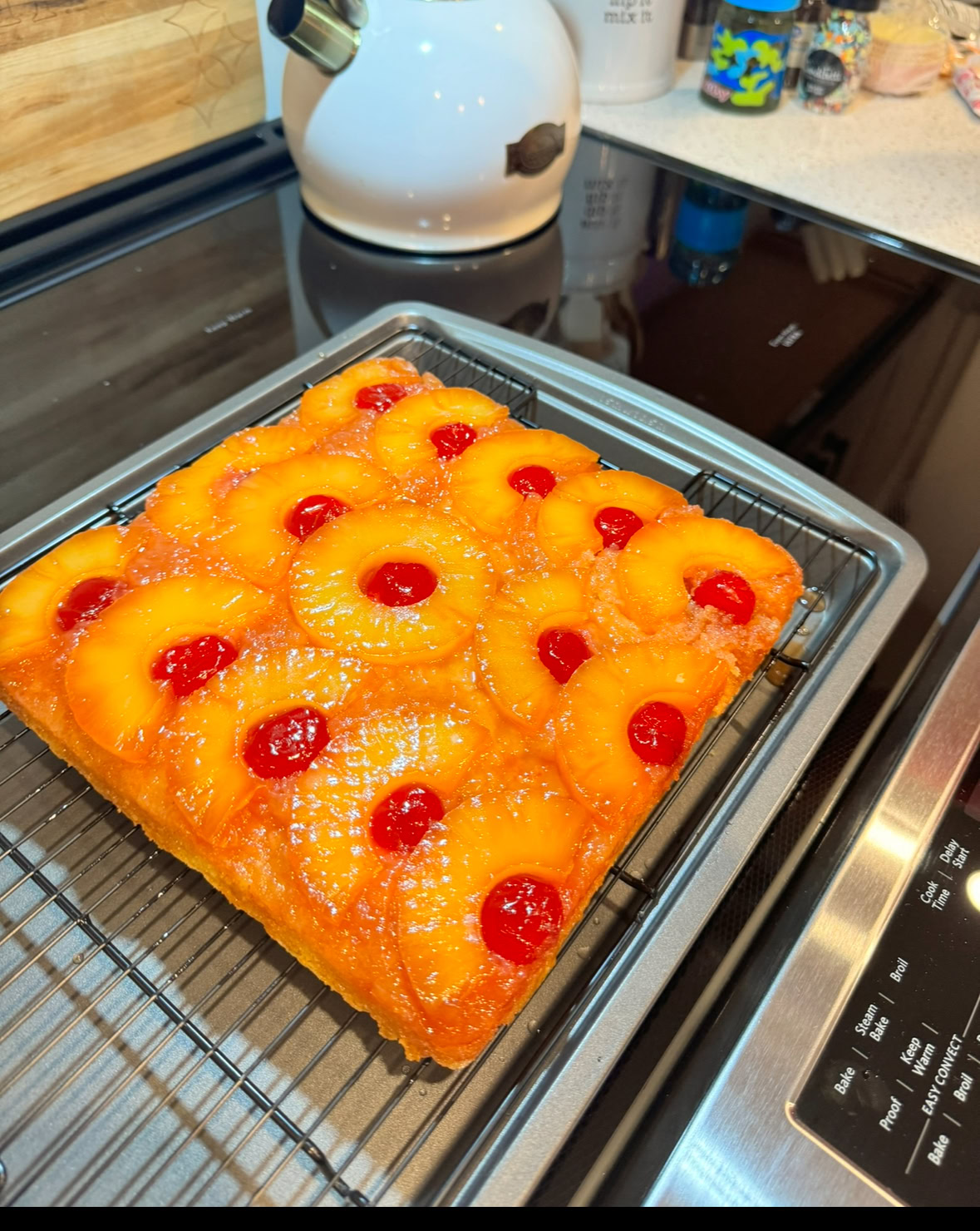 A square pineapple upside-down cake with pineapple rings and cherries on top, cooling on a wire rack on a kitchen counter.