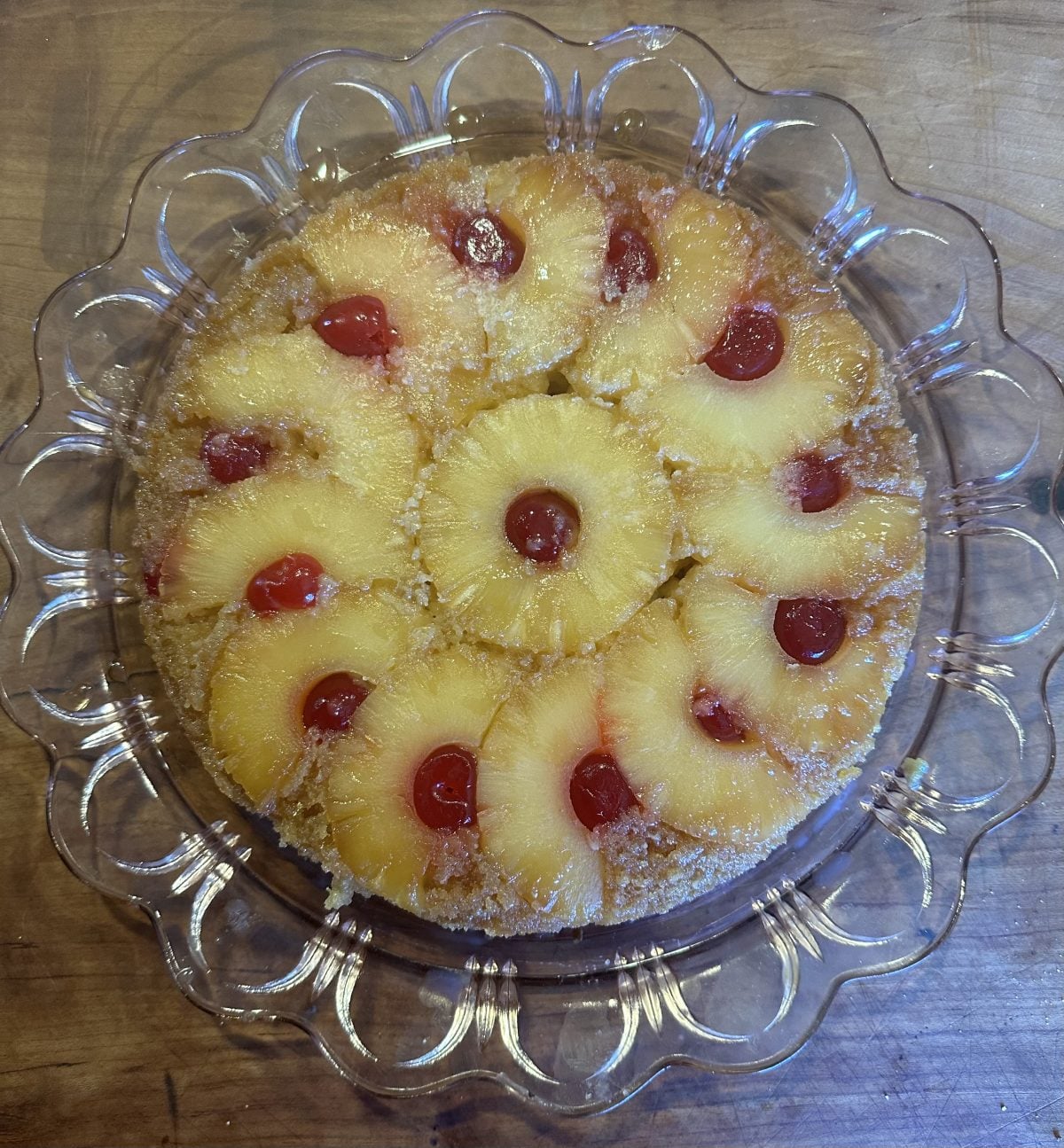 A pineapple upside-down cake with pineapple rings and maraschino cherries arranged on top, displayed on a clear glass plate.