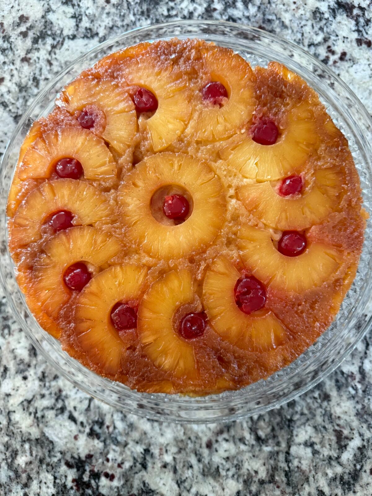 Pineapple upside-down cake with circular pineapple rings and cherries on top, displayed on a glass plate.