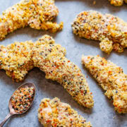Breaded chicken tenders coated with a variety of spices, lying on a light grey surface next to a spoonful of seasoning blend.