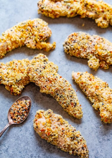 Breaded chicken tenders coated with a variety of spices, lying on a light grey surface next to a spoonful of seasoning blend.