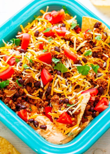 A colorful taco salad with ground beef, beans, shredded cheese, tomatoes, and cilantro in a blue dish, served with tortilla chips.
