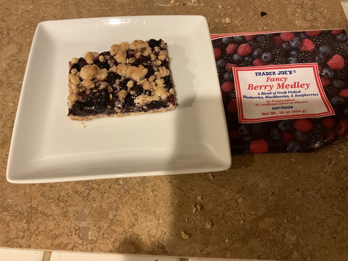 A berry crumble bar on a white plate next to a Trader Joe's Fancy Berry Medley package on a beige countertop.