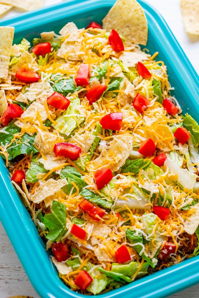 Layered taco salad with shredded cheese, lettuce, tomatoes, and tortilla chips in a blue dish.