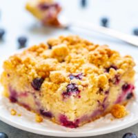 A slice of blueberry crumble cake on a white plate surrounded by fresh blueberries.
