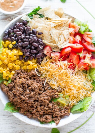 A colorful taco salad with layers of lettuce, ground beef, shredded cheese, black beans, corn, tomatoes, and crushed tortilla chips.