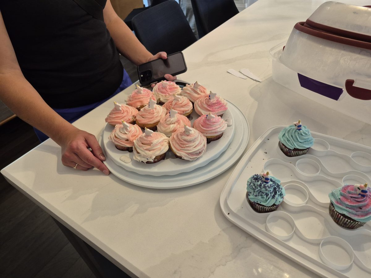A person holds a tray of cupcakes with pink and white frosting; next to it are three cupcakes with blue frosting on a white tray on a kitchen counter.