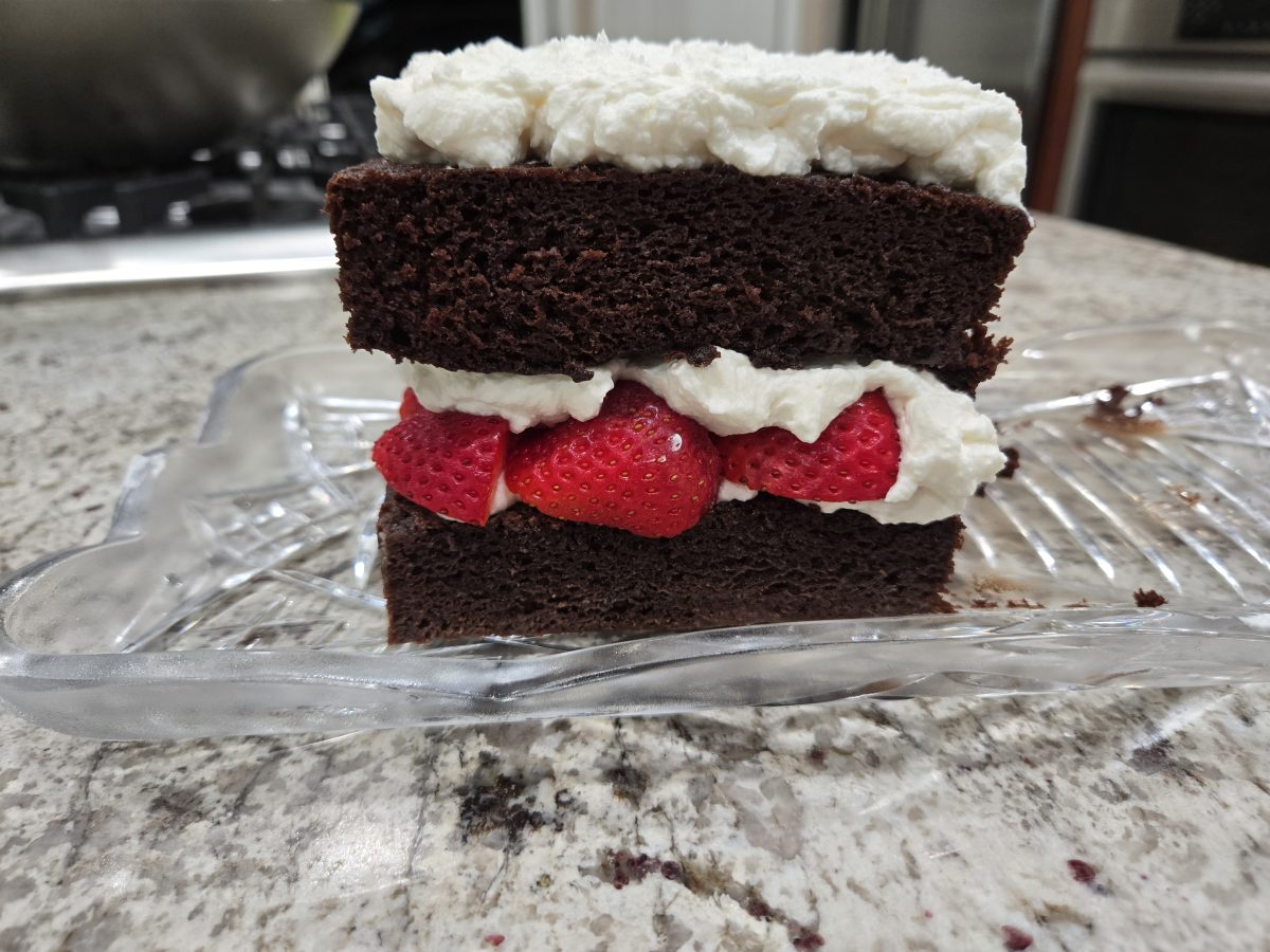 A slice of chocolate cake with a layer of strawberries and whipped cream in the middle and whipped cream on top, placed on a glass tray on a granite countertop.