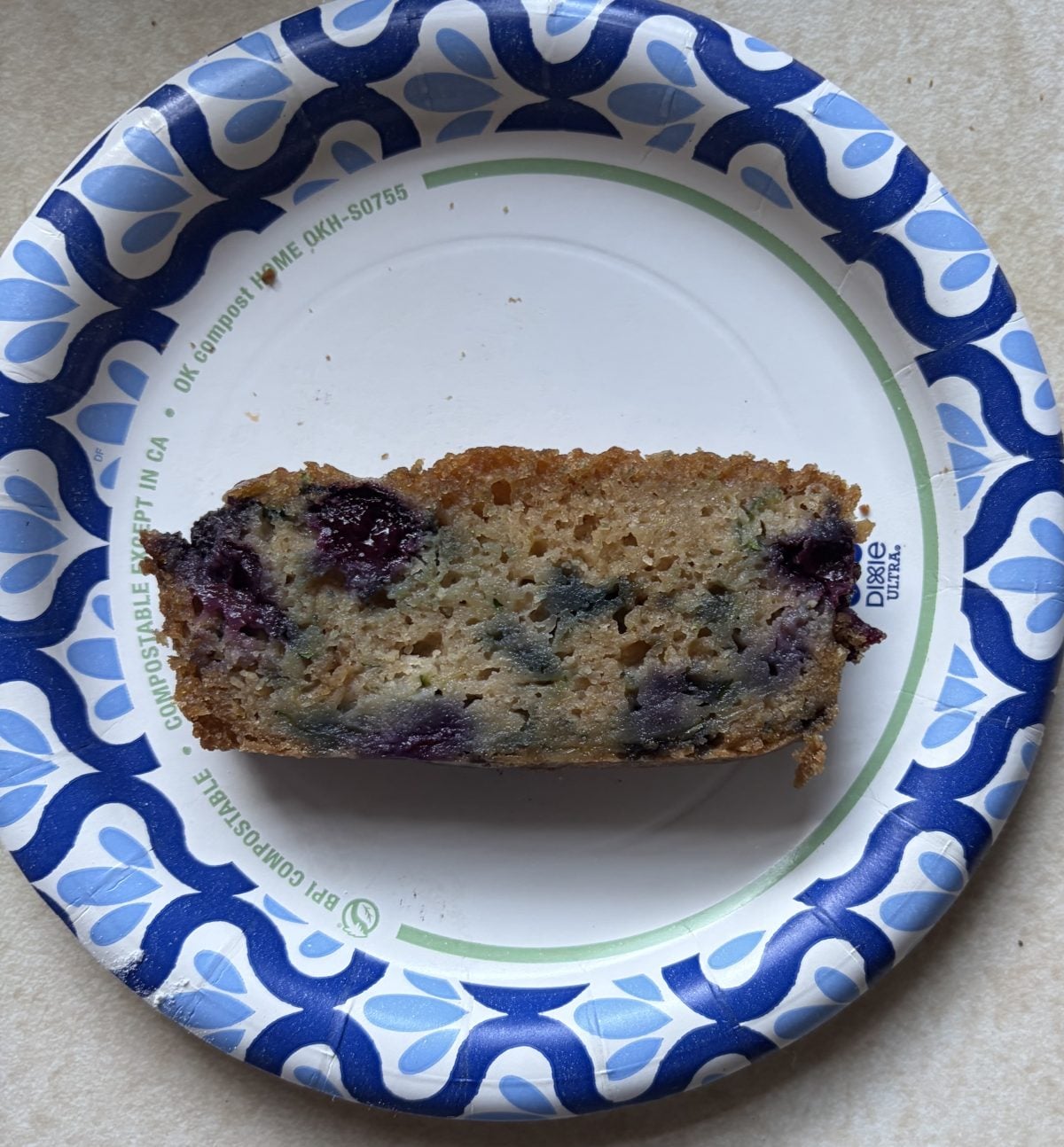 A slice of blueberry bread on a white paper plate with a blue geometric border, placed on a light-colored countertop.