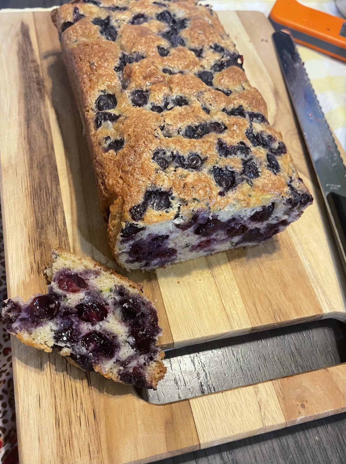 A loaf of blueberry bread sits on a wooden cutting board, with one slice cut and positioned in front, showing the blueberry-filled interior. A bread knife is nearby.