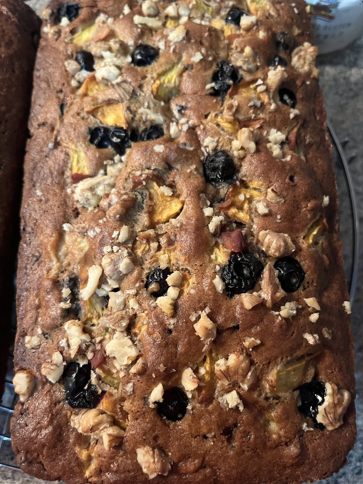 A loaf of homemade bread with visible chunks of fruit, blueberries, and chopped walnuts on top, cooling on a wire rack.