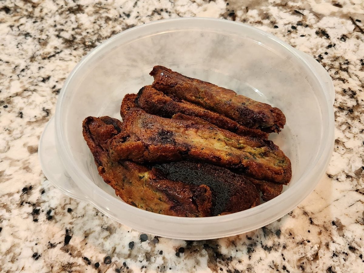 A plastic bowl containing several browned, possibly burnt, homemade vegetable patties or fritters sits on a marbled countertop.