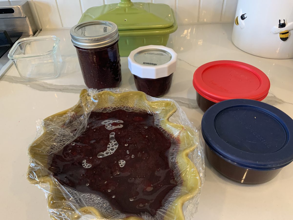 Several containers and jars filled with homemade berry jam are arranged on a kitchen counter, with one large bowl covered in plastic wrap.