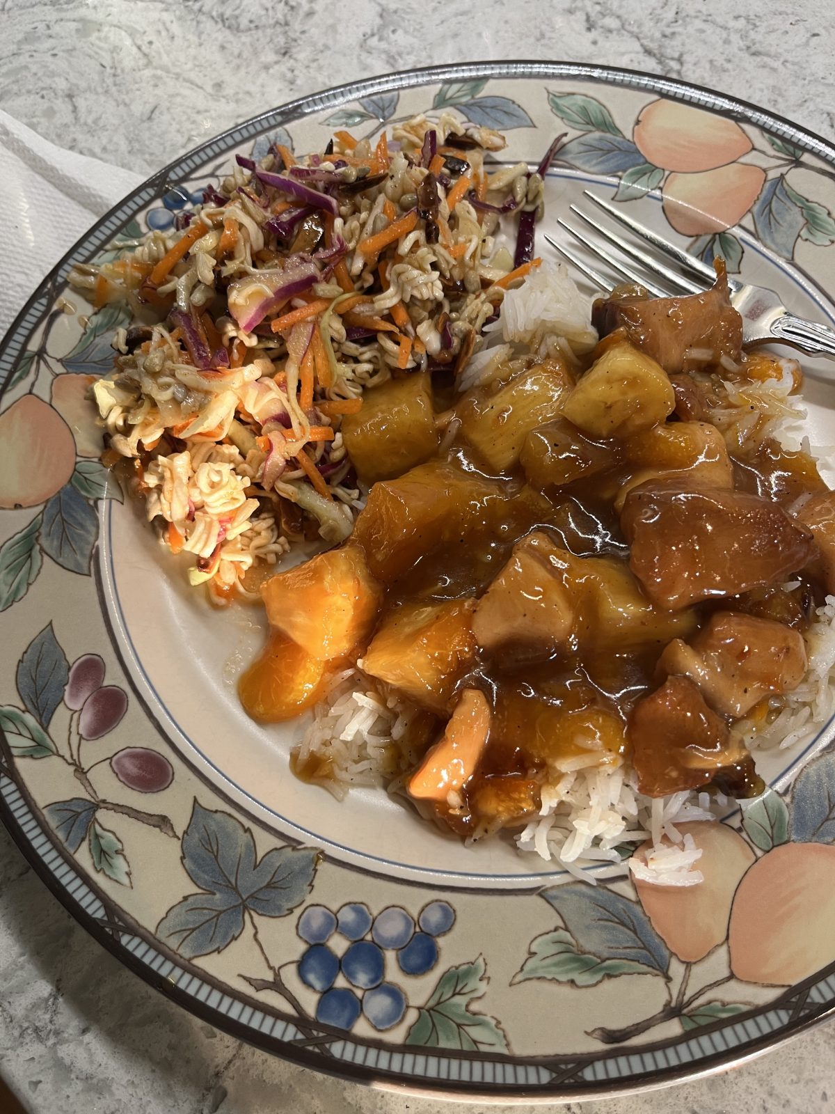 A plate with white rice topped with sweet and sour chicken and pineapple, served with a shredded cabbage and carrot slaw mixed with crunchy noodles.