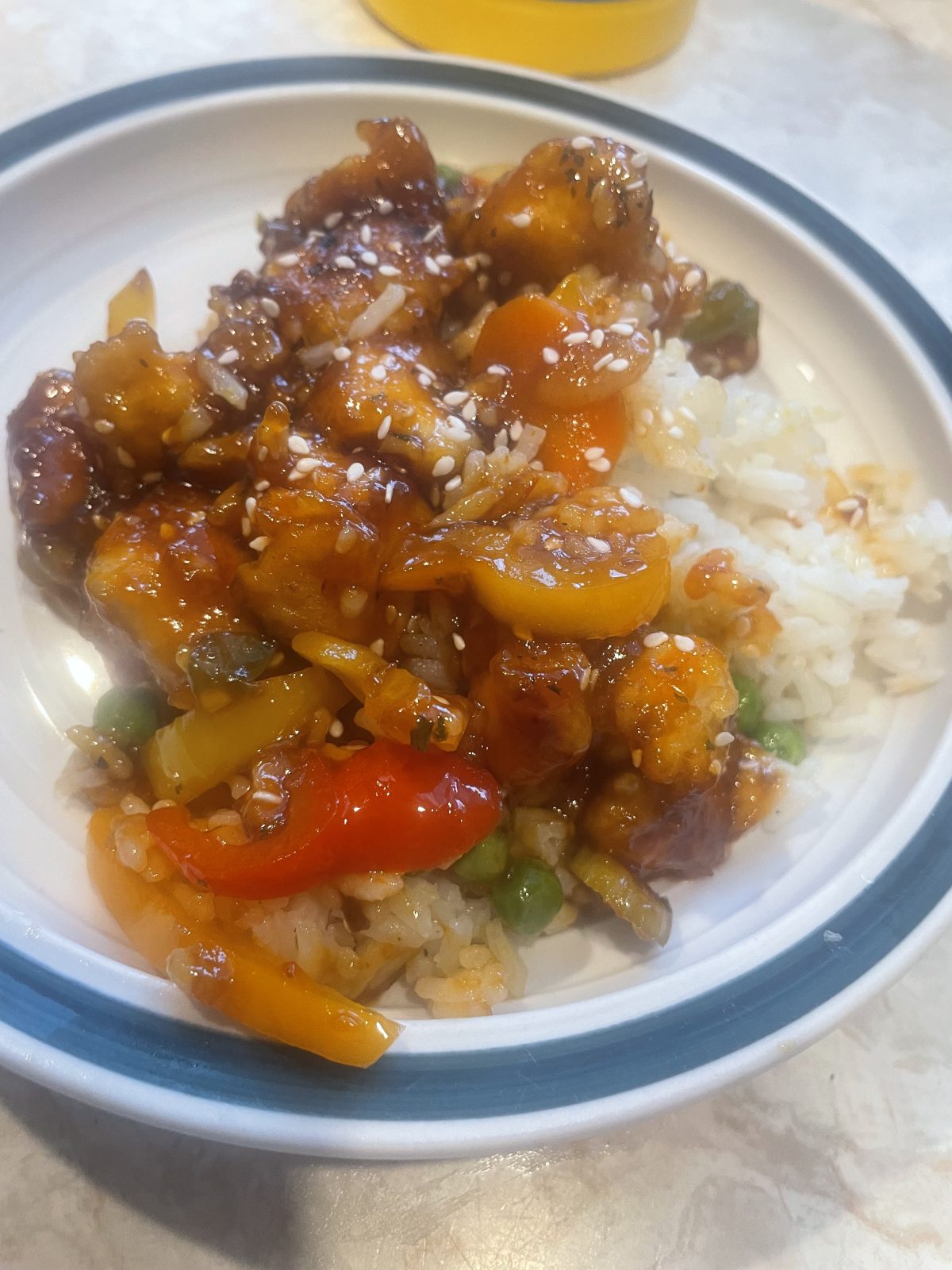 A plate of white rice topped with sweet and sour chicken, bell peppers, peas, and sesame seeds in a light sauce.