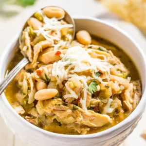 A bowl of white chicken chili topped with shredded cheese and garnished with peanuts, with a spoon inserted in the bowl.