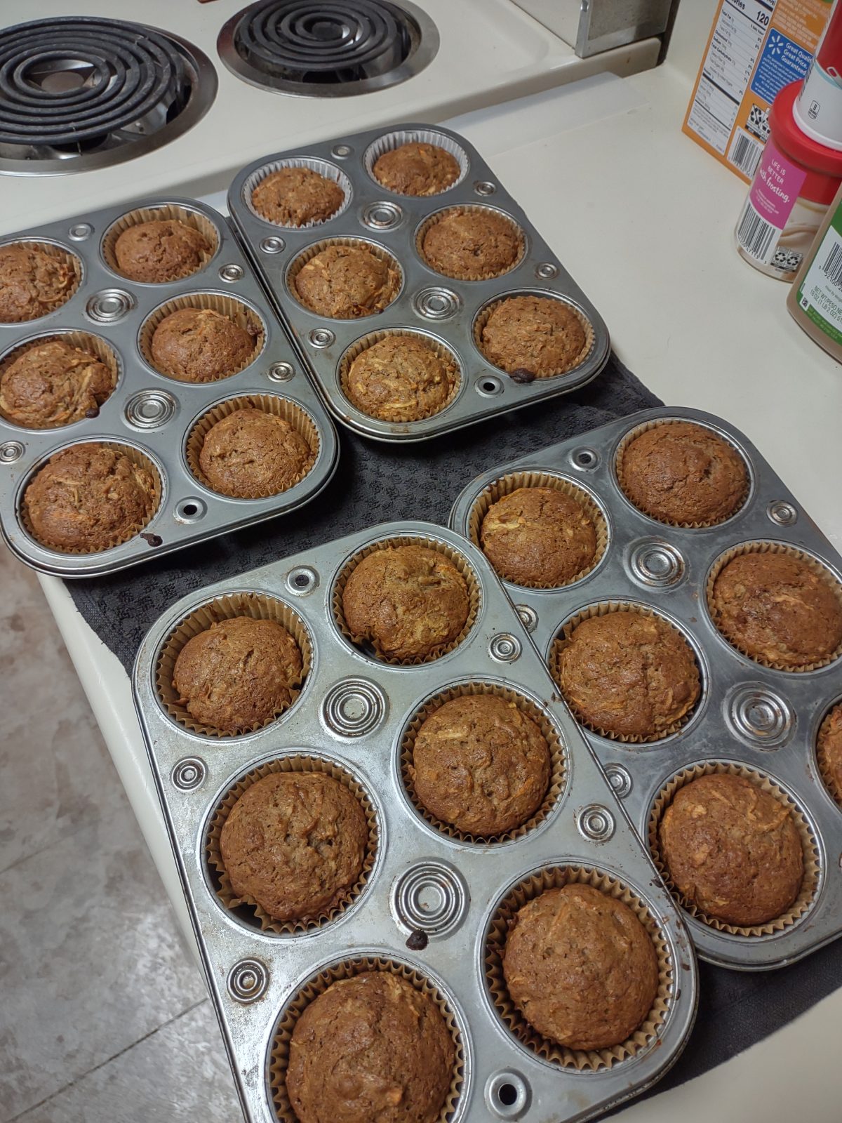 Four trays of freshly baked muffins cooling on a kitchen counter near a stove and spice containers.