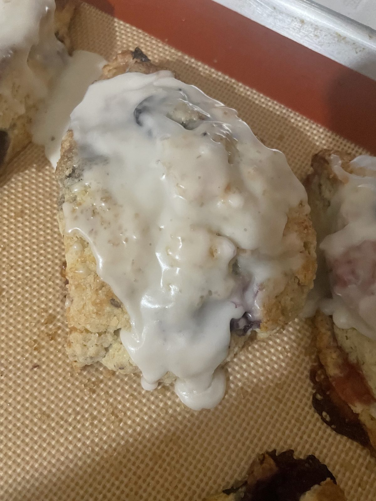 A triangular scone with visible berries is topped with a thick layer of white icing, resting on a silicone baking mat.