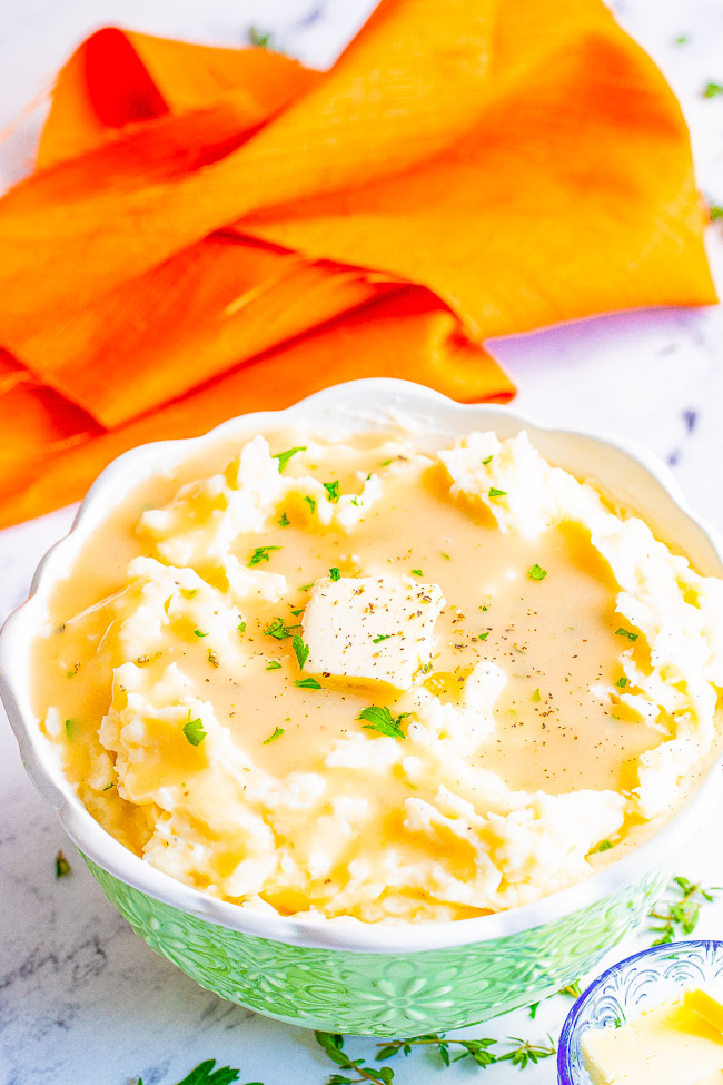 A large serving bowl of mashed potatoes from scratch topped with turkey gravy and a pat of butter. An orange towel lies in the background. 