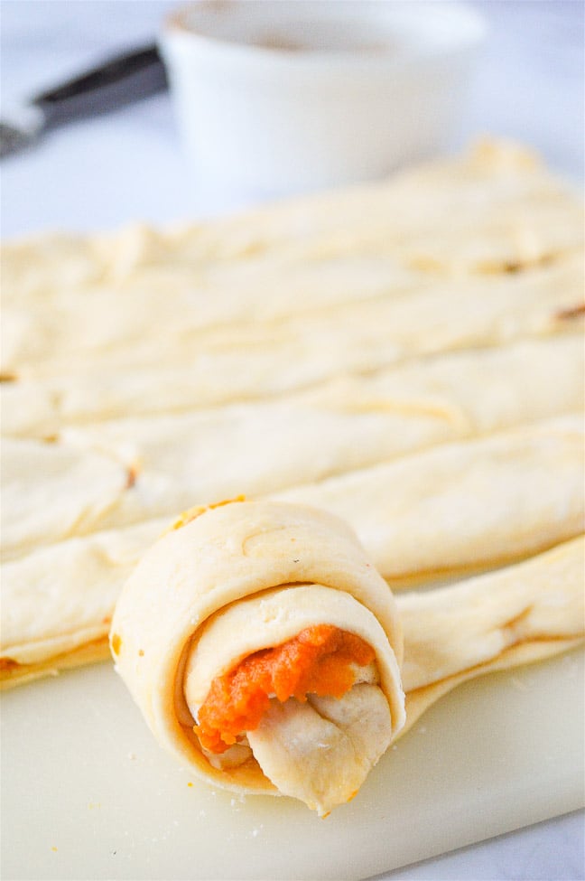 A rolled-up dough with a pumpkin filling on a cutting board, with more rolls and a bowl in the background.