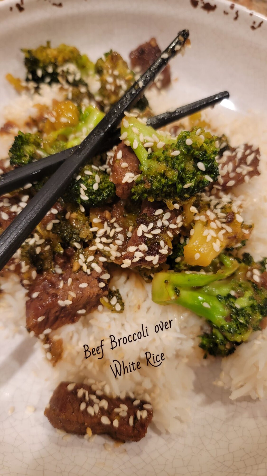 A dish of beef and broccoli with sesame seeds on white rice, served with black chopsticks.