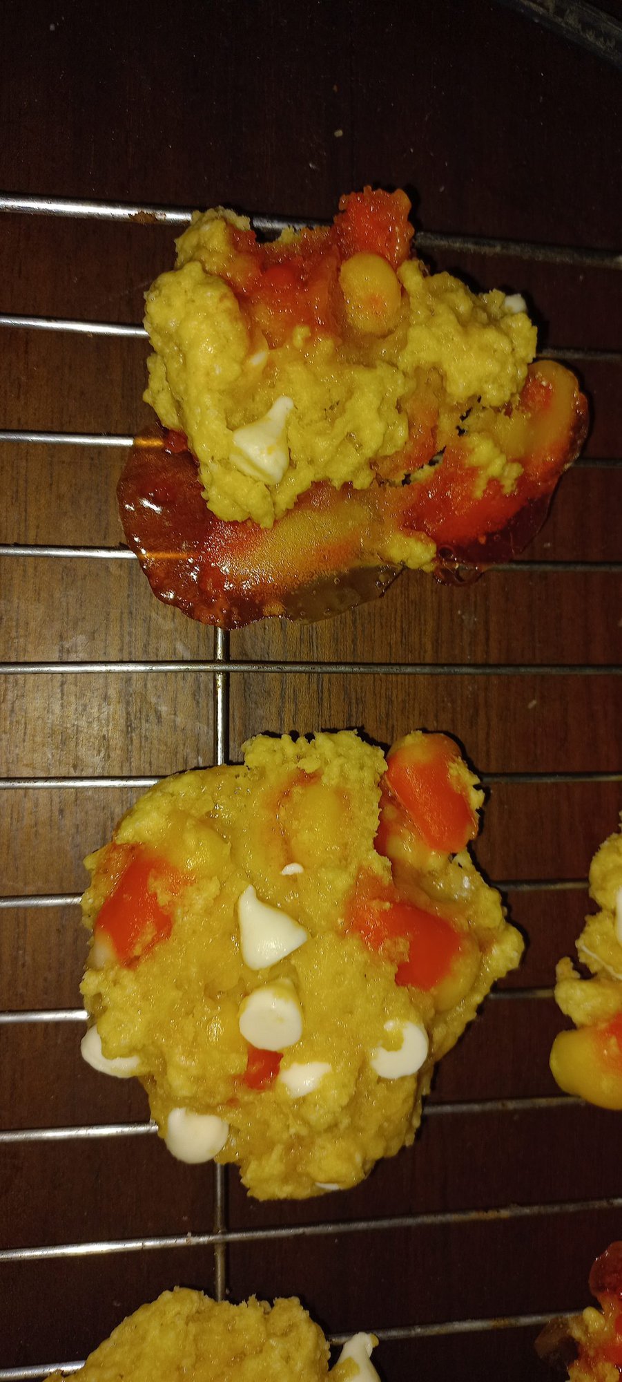 Two irregularly shaped cookies with visible white chocolate chips and orange pieces rest on a cooling rack over a wooden surface.