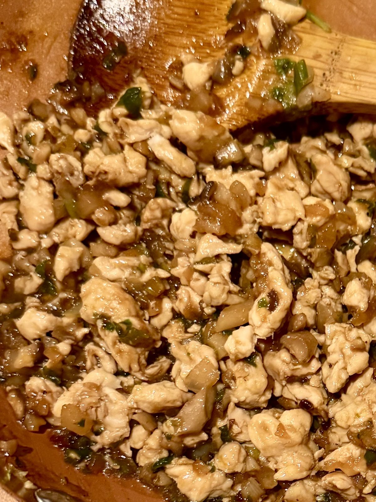 Close-up of chopped chicken sautéed with onions, herbs, and sauce in a pan, with a wooden spoon partially visible.
