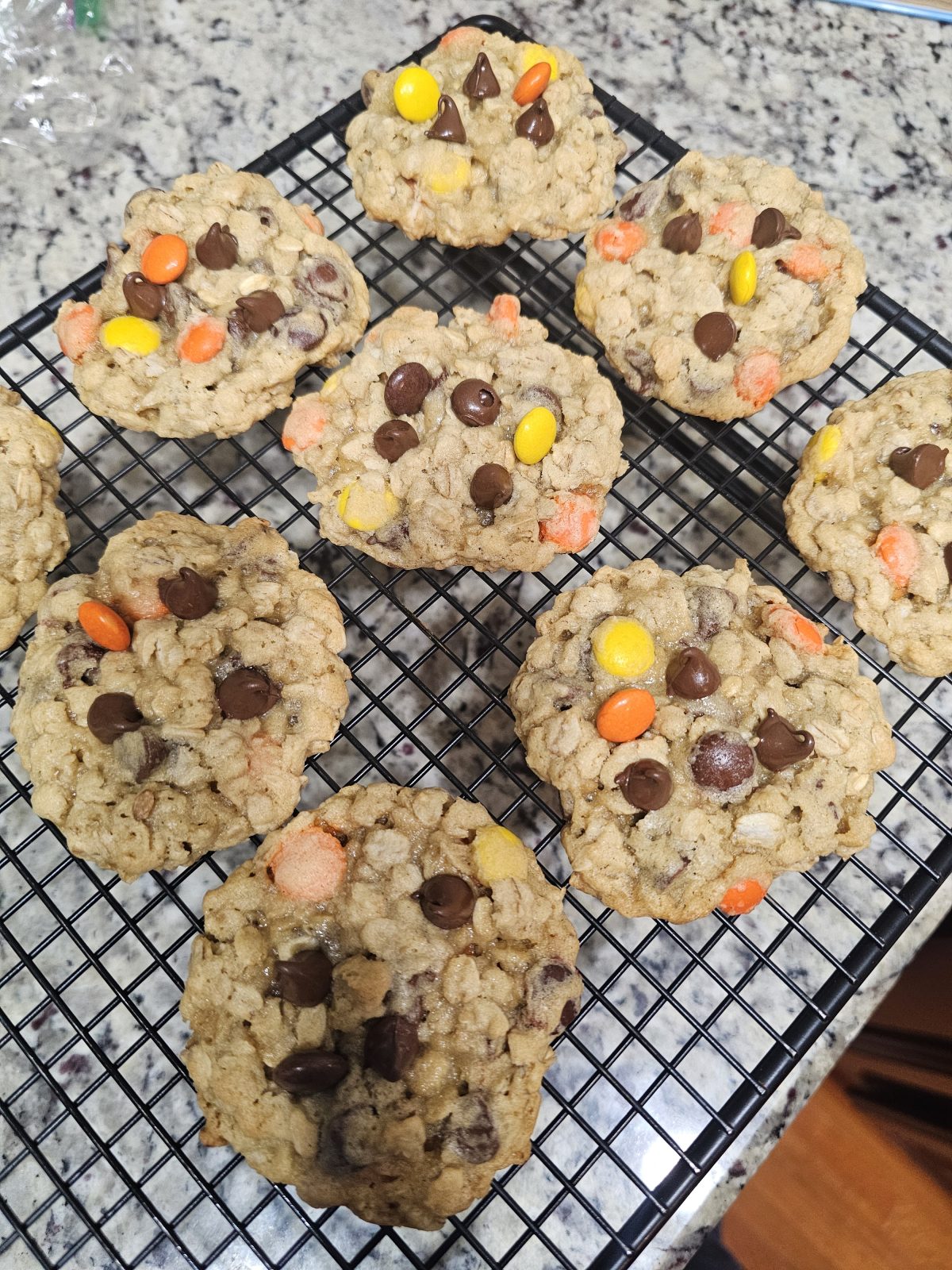 Nine freshly baked cookies with chocolate chips and colorful candy pieces are cooling on a black wire rack on a granite countertop.