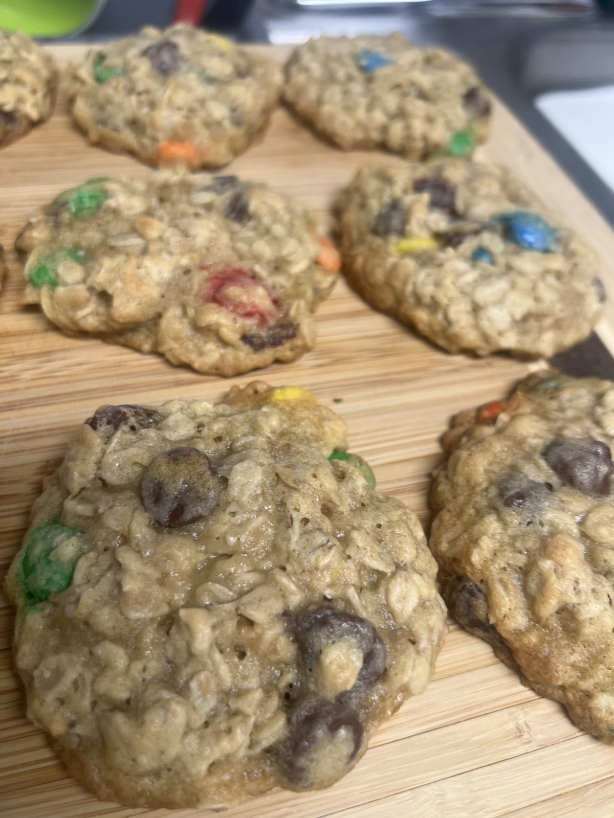 A close-up of homemade oatmeal cookies with chocolate chips and colorful candy pieces on a wooden cutting board.