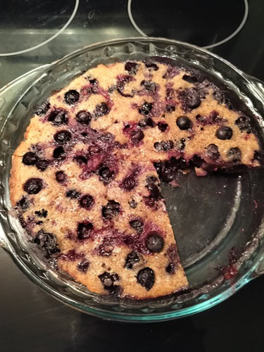 A round glass baking dish with a blueberry cake, one slice missing, sits on a stovetop.