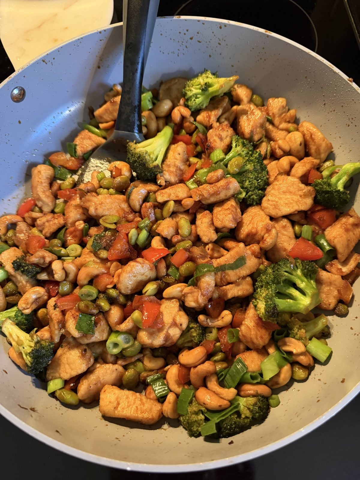 A pan filled with a chicken stir-fry, including pieces of chicken, broccoli, bell peppers, green onions, cashews, and snap peas, with a serving spoon inside.