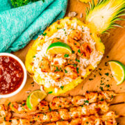 Pineapple Teriyaki Chicken Skewers on a cutting board next to a pineapple half filled with white rice and chicken pieces.