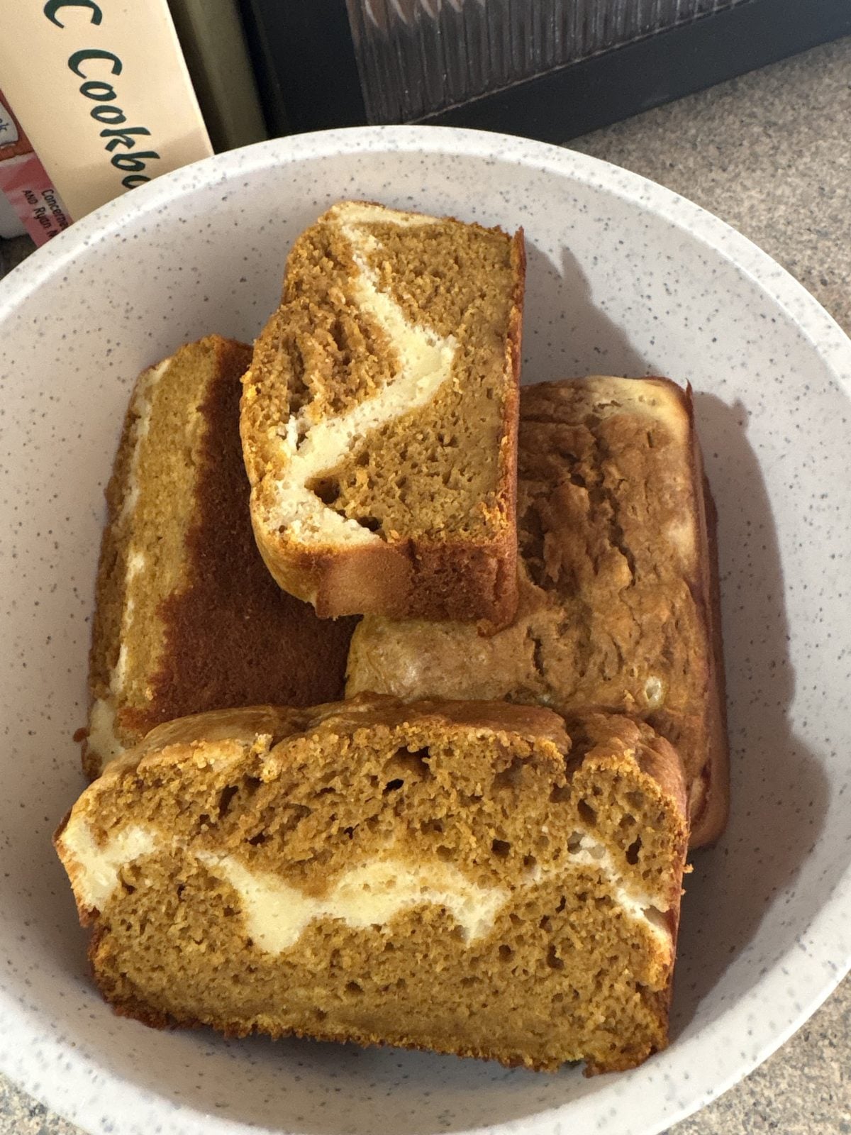A white speckled bowl contains several slices of pumpkin bread with a visible cream cheese swirl on a kitchen countertop.